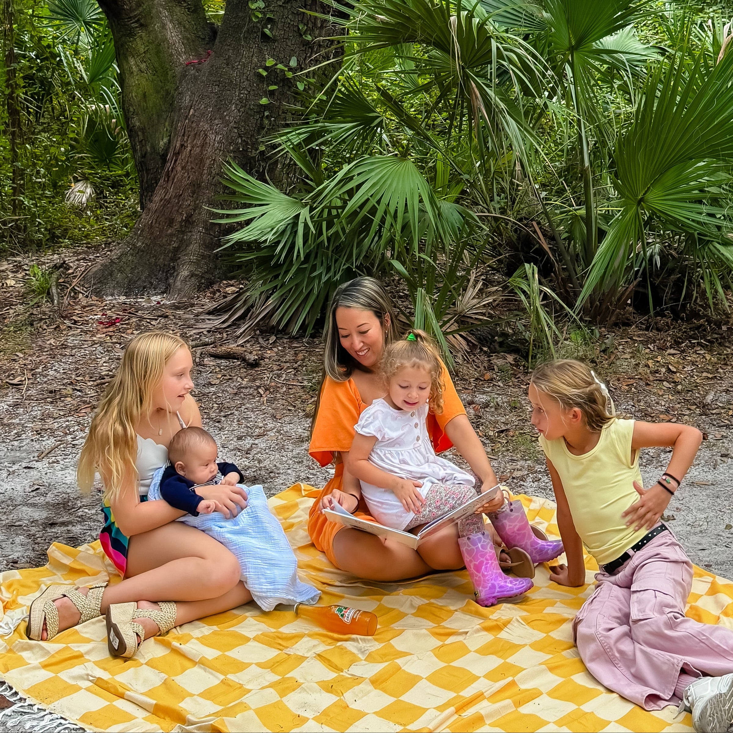 Four children sitting on a yellow patterned blanket under a large tree with greenery reading a children's book with the author